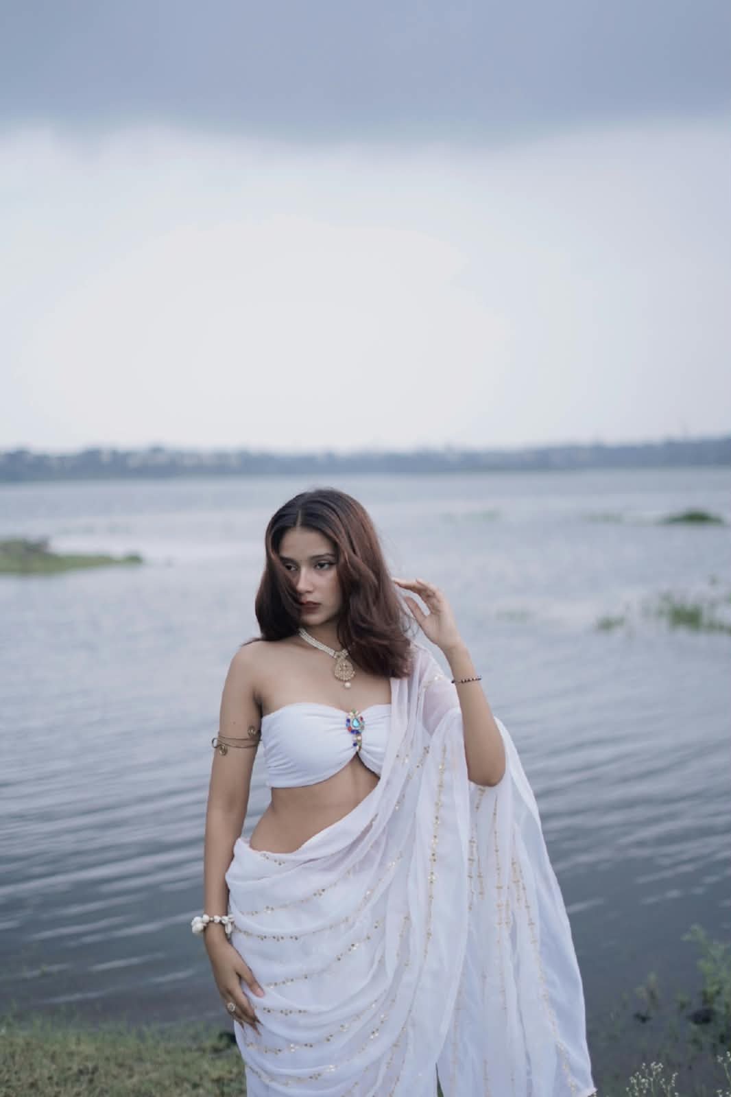 Janvi Saxena in a white saree against a cloudy lakeside sky